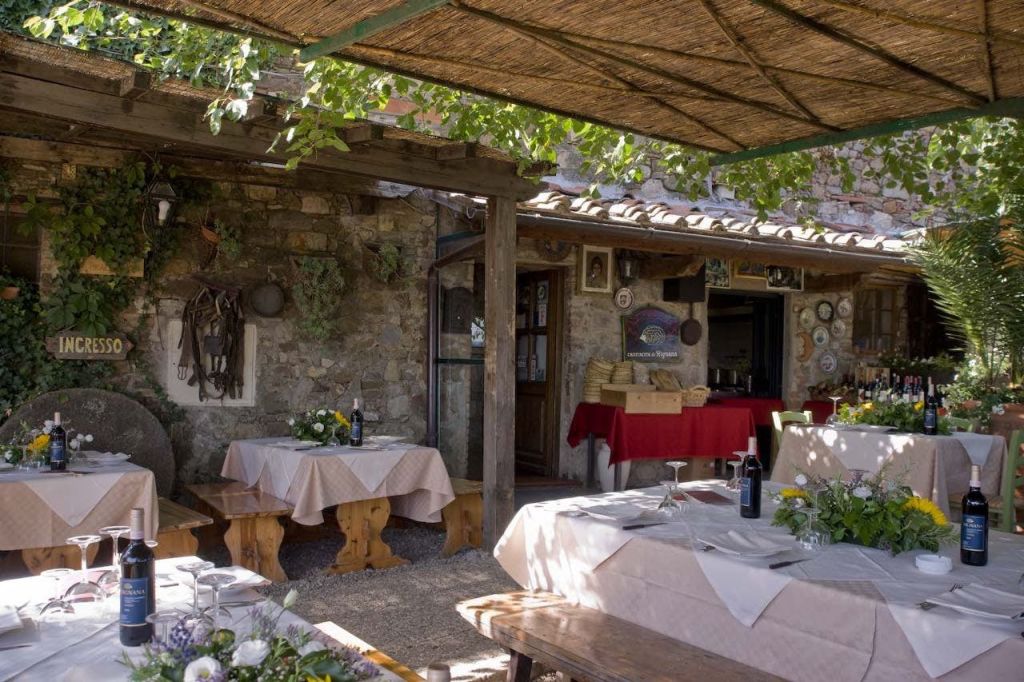 녹음이 우거진 나무 퍼걸러로 덮인 돌담이 있는 소박한 레스토랑의 야외 좌석. 테이블에는 침구가 놓여 있습니다.