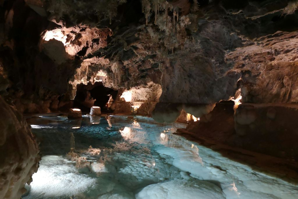 밝게 빛나는 종유석과 석순이 잔잔하고 맑은 물에 비치는 고요한 지하 동굴 장면.