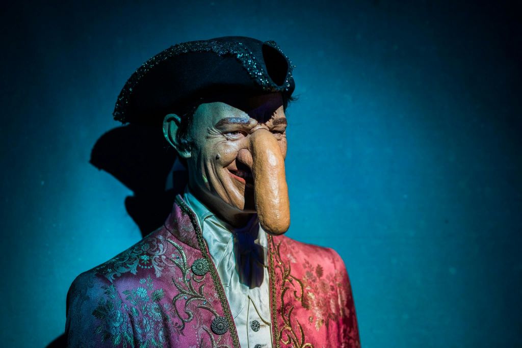 커다랗고 과장된 코와 파란색 배경에 검은색 삼각 모자가 달린 섬세하고 화려한 의상을 입은 인물.