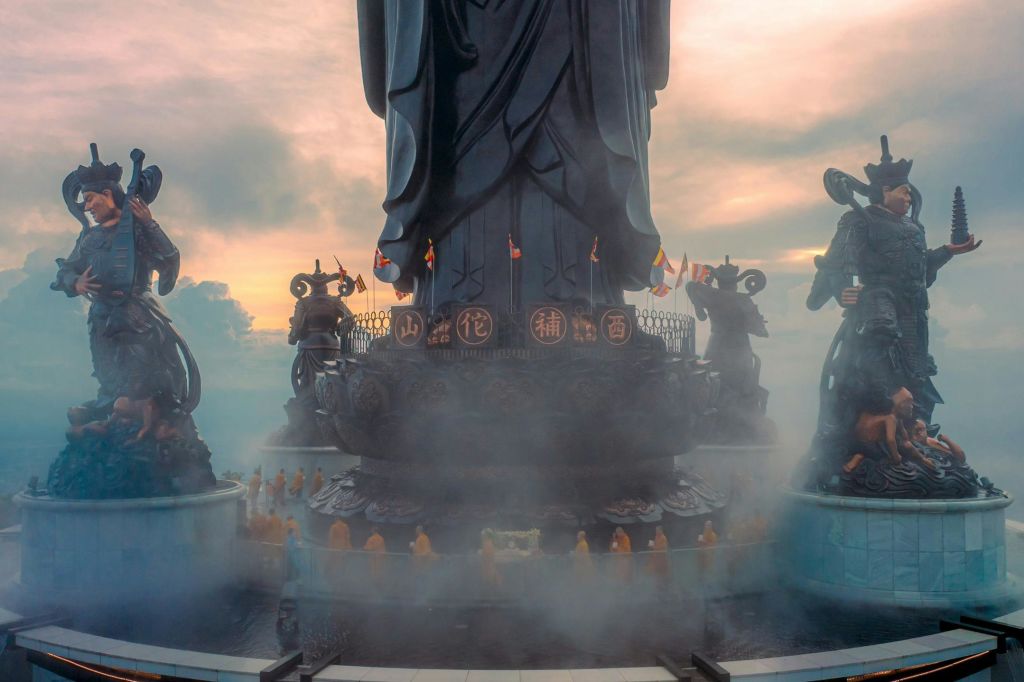 안개로 둘러싸인 주위에 작은 조각상이 있는 큰 조각상. 주황색 가운을 입은 수도승들이 기지 주변에 배치되었습니다. 석양에 흐린 하늘.