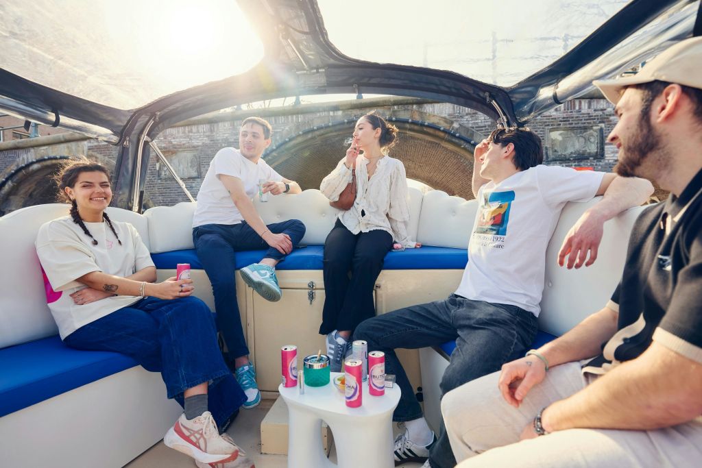 작은 테이블 위, 캐노피 아래, 햇살이 내리쬐는 하늘 아래에서 음료를 마시며 보트 갑판에 앉아 있는 네 명의 사람들.