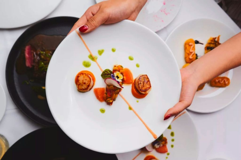 예술적으로 배열된 가리비, 장식, 얇은 소스 라인이 있는 흰색 접시를 들고 있는 손. 배경에 다른 플레이트가 보입니다.