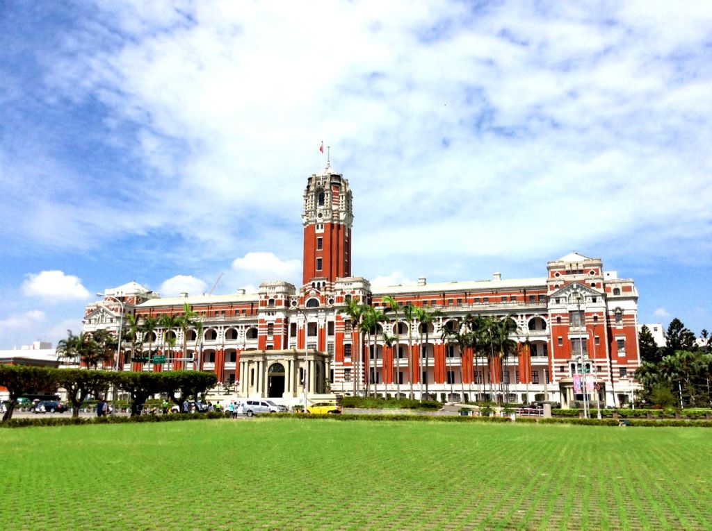 부분적으로 흐린 하늘 아래 녹음으로 둘러싸인 높은 시계탑이 있는 웅장한 붉은색과 흰색의 식민지 양식 건물입니다.