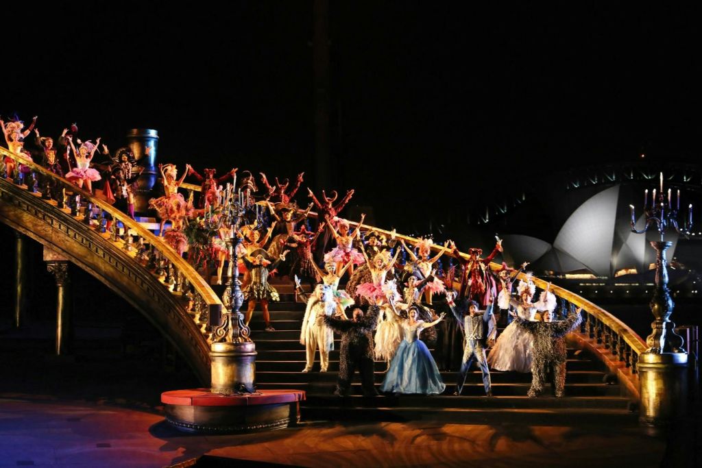 어두운 배경을 배경으로 한 웅장한 계단에서 의상을 입은 많은 배우들의 연극 공연이 펼쳐집니다.