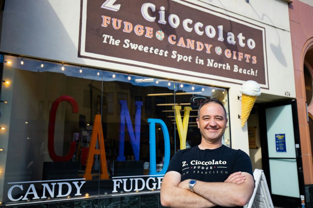 한 사람이 "Z. Cioccolato" 라는 이름의 퍼지와 사탕 가게 앞에 "노스 비치에서 가장 달콤한 장소" 라는 팻말을 들고 팔짱을 끼고 웃으며 서 있습니다!