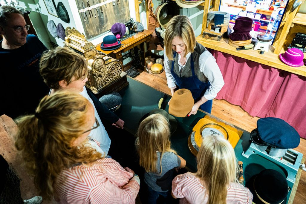 어린이를 포함한 한 무리의 사람들이 빈티지 상점에서 모자 제작 시연을 하는 여성 주위에 모입니다.