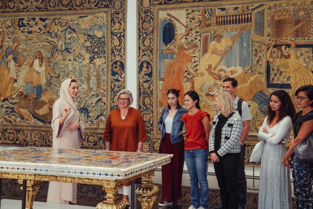 한 무리의 사람들이 미술관에 서서 정교한 디자인과 역사적인 장면이 있는 크고 화려한 태피스트리를 관찰하고 있습니다.