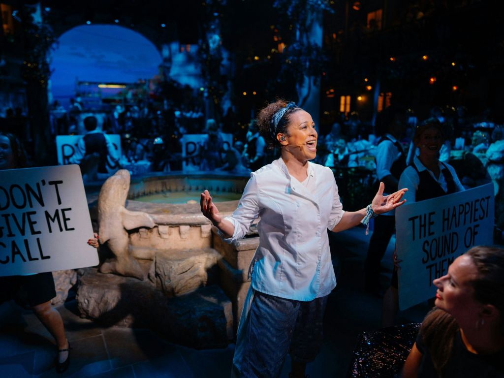 흰색 셔츠를 입은 여성이 분수와 관객을 배경으로 조명이 어두운 장소에서 열정적인 공연을 펼치고 있습니다.