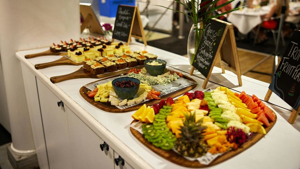 테이블 위에 다양한 과일, 치즈, 디저트가 놓여 있습니다. 작은 칠판 표지판에는 사용 가능한 식품이 나열되어 있습니다.