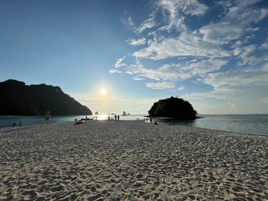 섬, 잔잔한 바다, 부분적으로 흐린 하늘 아래 석양이 지는 모래 해변에서 사람들이 걷고 휴식을 취하고 있습니다.