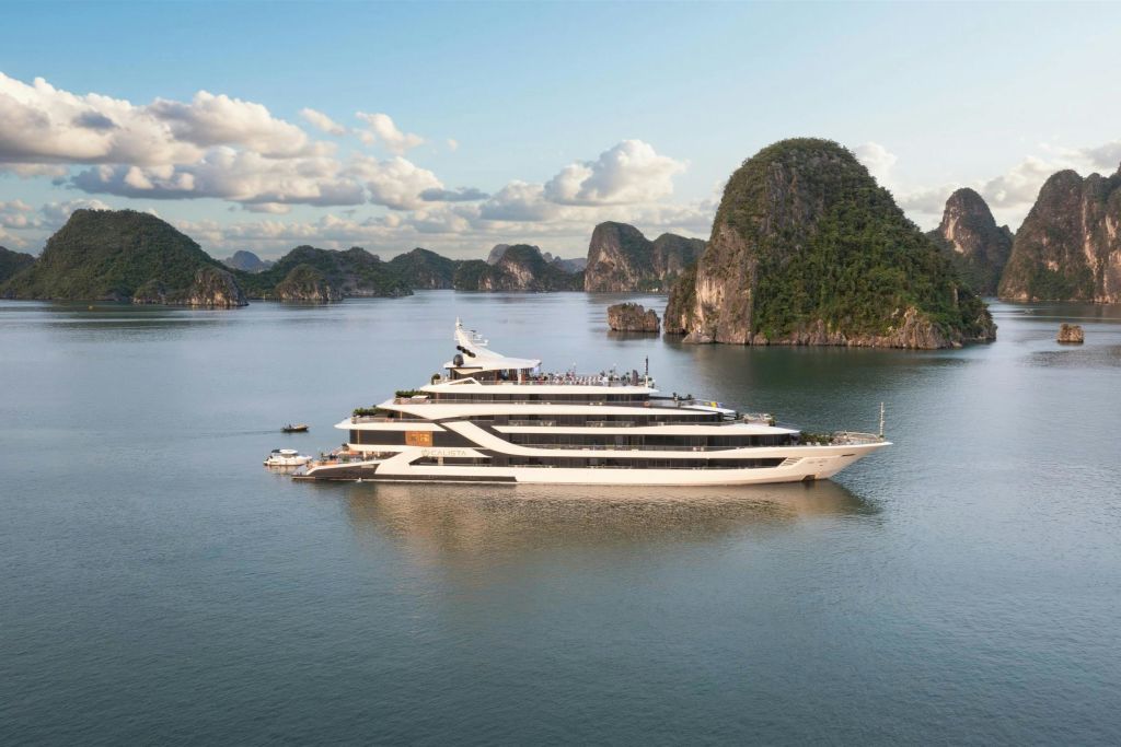 부분적으로 흐린 하늘 아래 녹색의 바위 섬으로 둘러싸인 잔잔한 물 위에 여러 개의 데크가 있는 커다란 흰색 요트가 떠 있습니다.