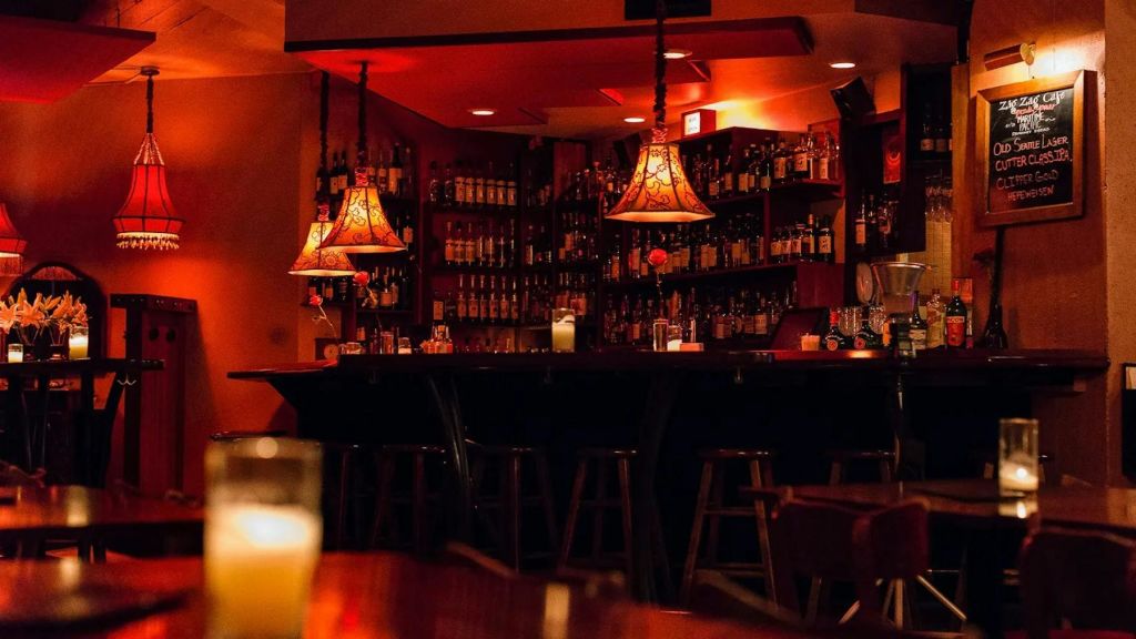 어두운 조명, 화려한 걸이식 램프, 병이 구비된 선반, 나무 바 의자가 있는 아늑한 바.