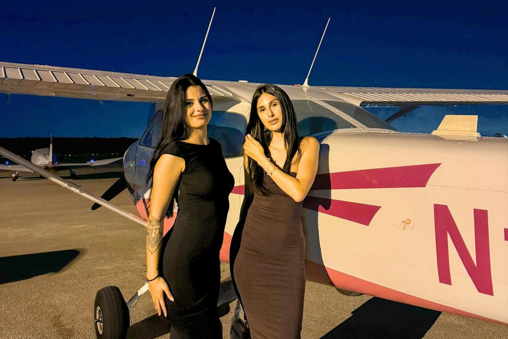 검은 드레스를 입은 두 명의 여성이 해질 무렵 활주로에서 작은 비행기 앞에 서 있습니다.