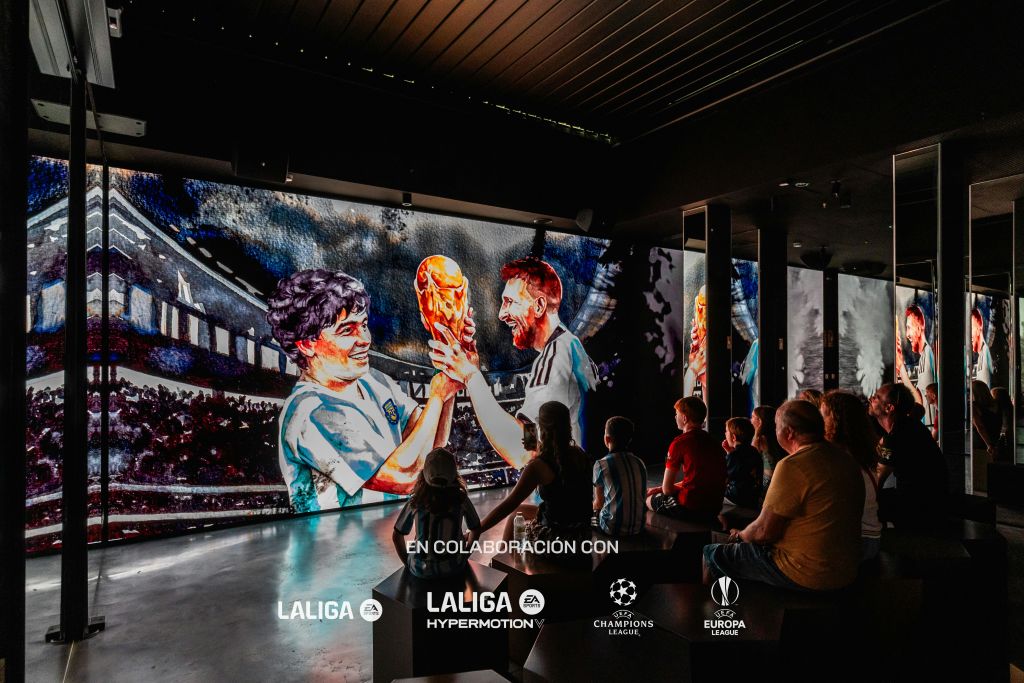 어두운 방에 앉아 두 명의 축구 선수가 트로피를 들고 있는 애니메이션 장면을 보여주는 대형 스크린을 보고 있는 사람들.