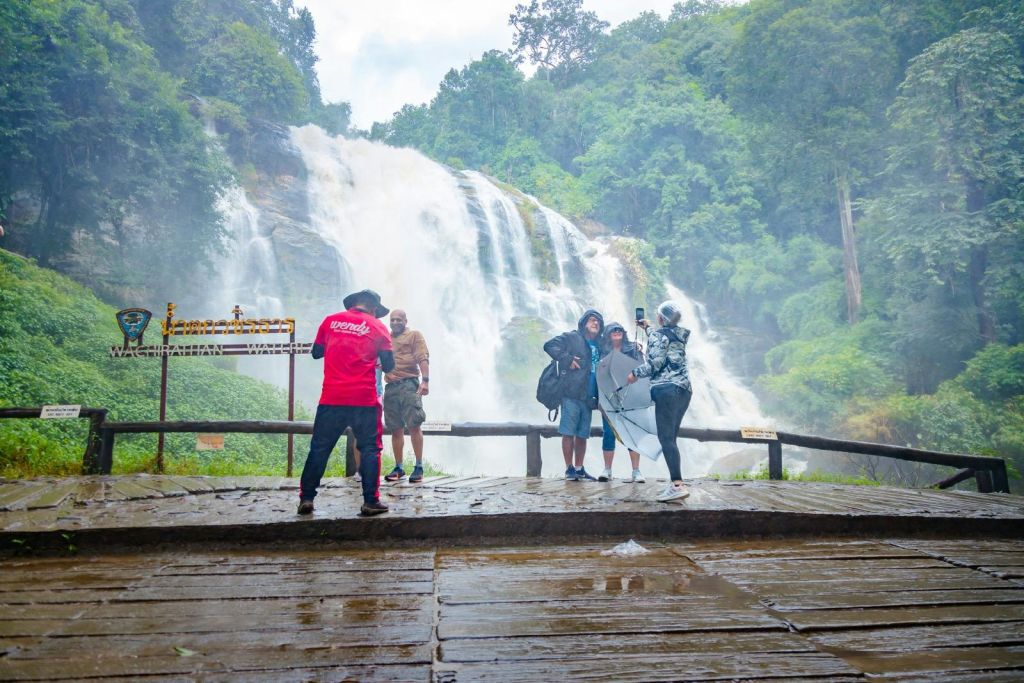 울창한 녹음으로 둘러싸인 커다란 폭포 앞에서 사람들이 젖은 나무 전망대에서 사진을 찍으며 포즈를 취하고 있습니다.