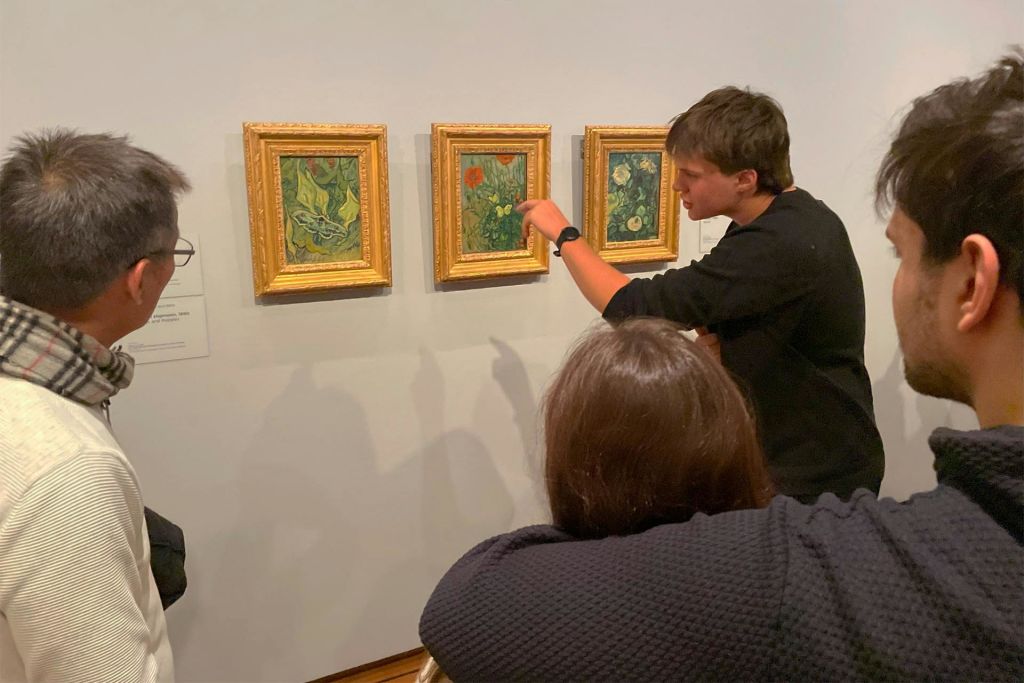 한 사람이 벽에 걸린 액자 그림을 향해 제스처를 취하는 동시에 갤러리에서 두 사람에게 말을 건넨다.