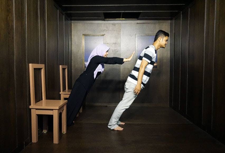 착각의 방에 있는 두 사람: 여자가 의자에 기대고 있는 동안 남자는 불가능한 각도로 앞으로 기울어져 있는 것처럼 보입니다.