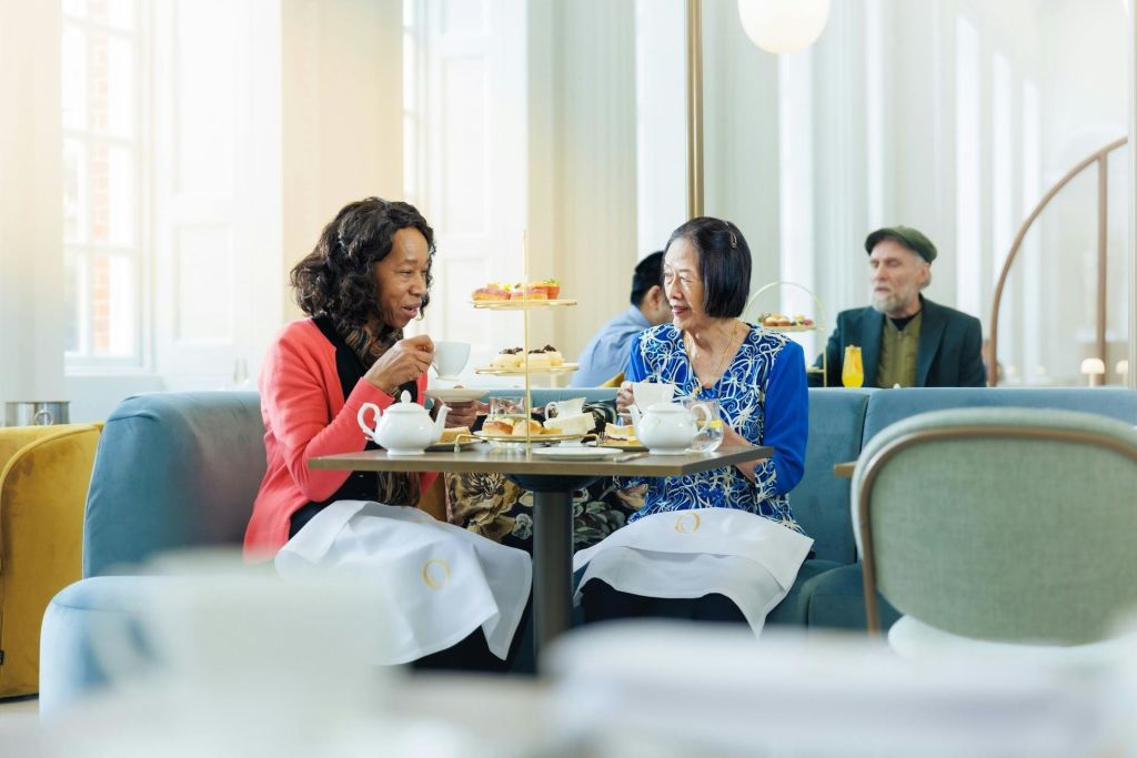 두 여성은 밝고 우아한 방에서 페이스트리와 컵을 층층이 쌓아 놓은 테이블에서 애프터눈 티를 즐깁니다.