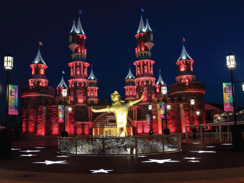 야간에 포탑이 있는 조명이 켜진 성으로, 팔을 앞으로 쭉 뻗은 모습의 중앙 황금 동상이 있습니다. 바닥에 흰색 별 무늬가 있습니다.