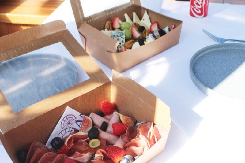 테이블 위에 놓인 두 개의 열린 판지 상자에는 다양한 고기, 치즈, 과일, 견과류가 들어 있습니다. 코카콜라 캔이 부분적으로 보입니다.