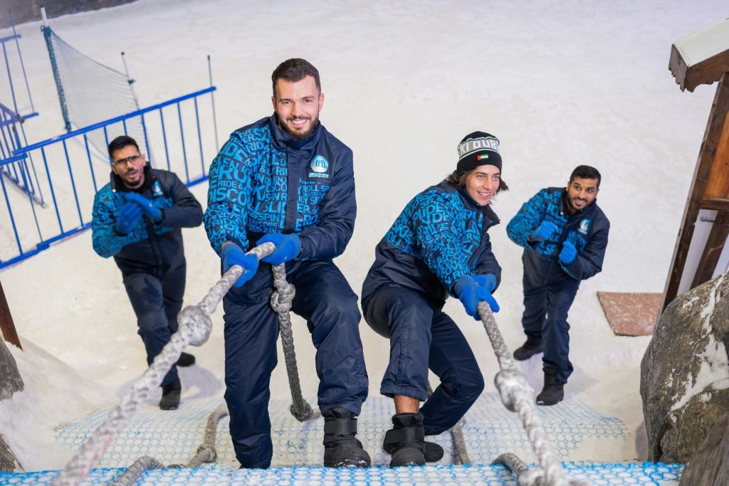 겨울 장비를 착용한 네 명이 실내 눈 환경에서 밧줄 그물을 타고 미소를 지으며 카메라를 바라보고 있습니다.