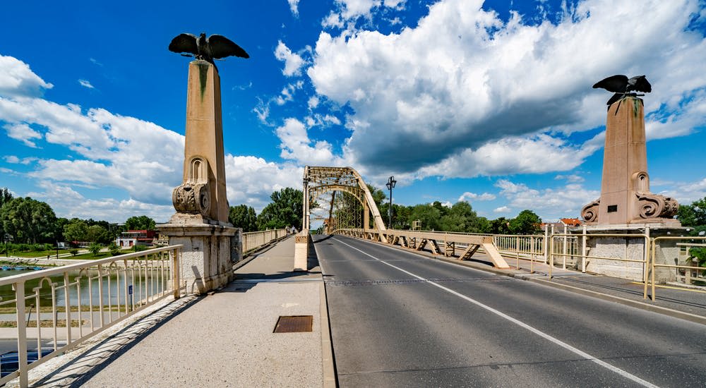 구름이 흩어져 있는 푸른 하늘 아래, 커다란 새상 기둥이 있는 텅 빈 도로 다리. 나무가 배경으로 보입니다.