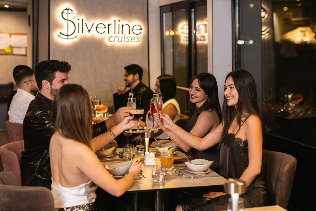 창문과 간판이 보이는 따뜻한 실내 환경에 앉아 토스트로 안경을 들어 올리는 식탁의 사람들.