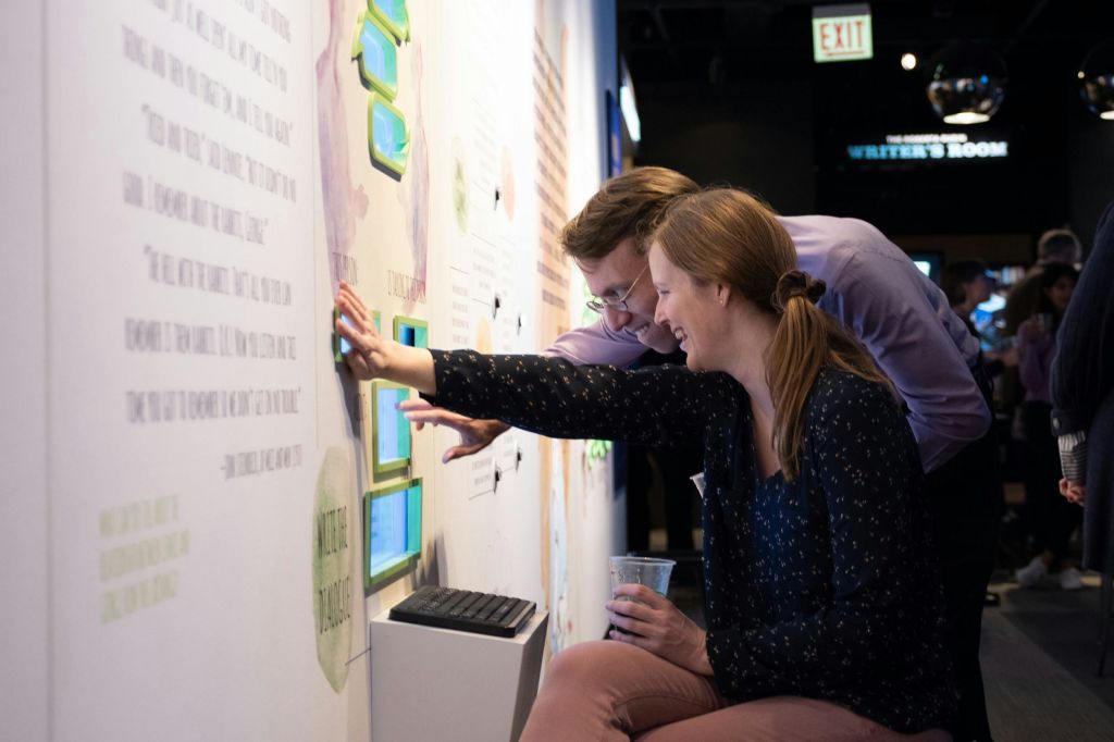 두 사람이 박물관 벽에 설치된 대화형 터치스크린 디스플레이에서 화면을 가리키며 미소를 짓고 있습니다.