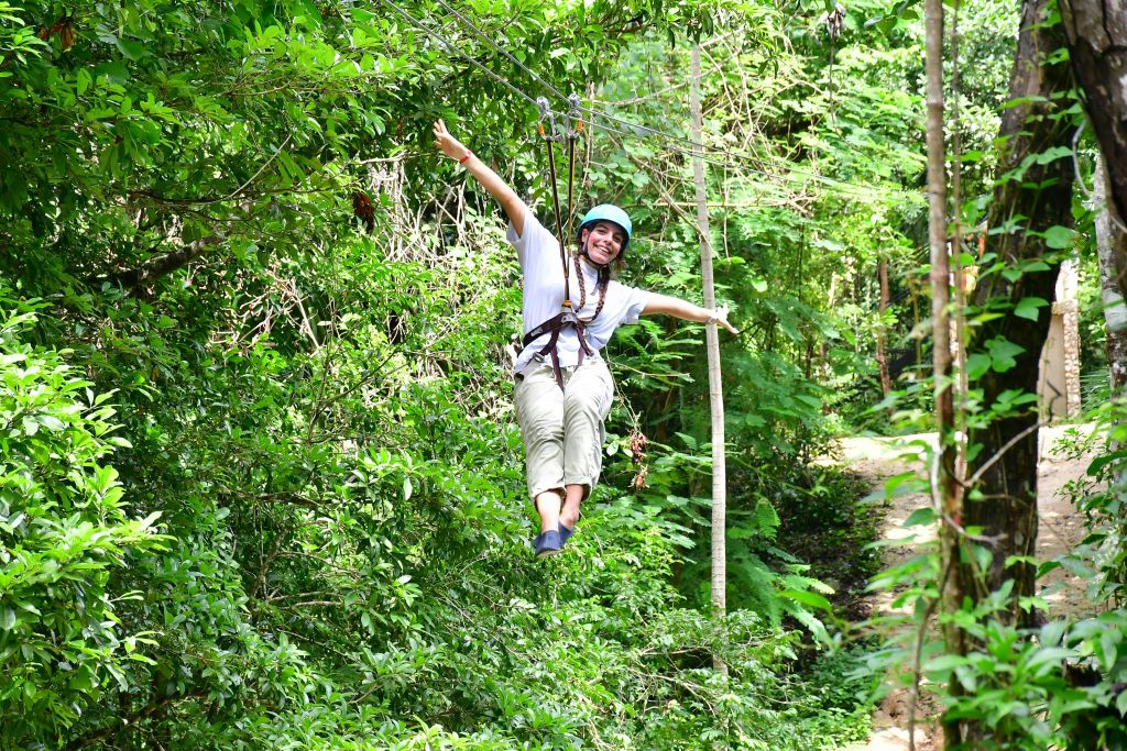 울창한 푸른 숲을 가로질러 미소를 짓고 집라인을 켠 사람은 안전 하네스와 헬멧을 착용하고 있습니다.