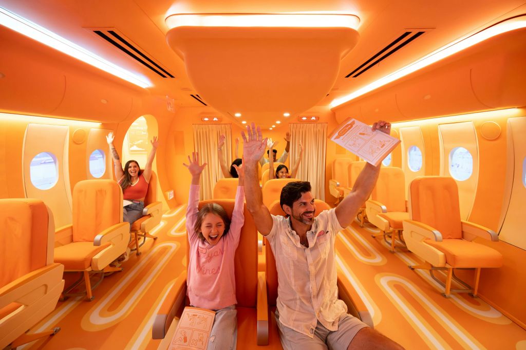 항공사 스타일의 좌석이 있는 밝은 조명, 주황색 테마의 방에 앉아 팔을 들고 활동 시트를 들고 있는 사람들.