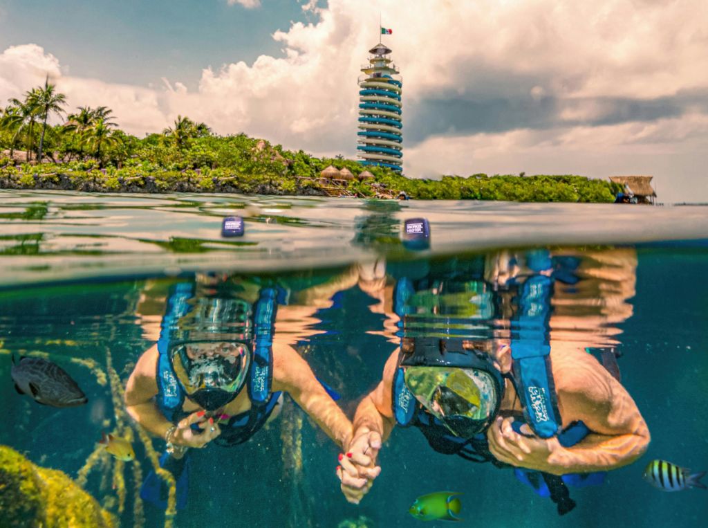 흐린 하늘을 배경으로 복층 전망대가 있는 푸른 섬 근처에서 두 명의 스노클러가 물속에서 스노클링을 즐깁니다.