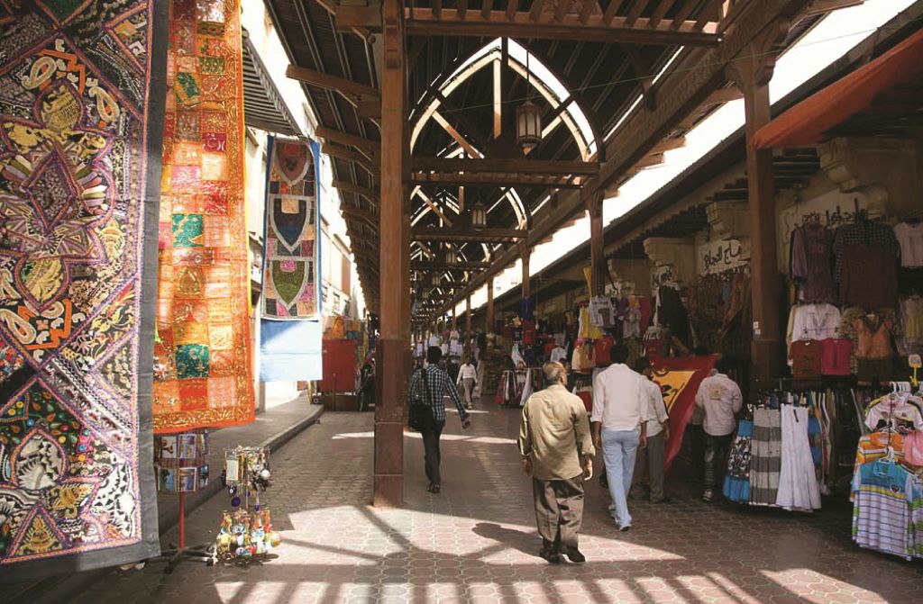 사람들은 나무 아치 아래 화려한 직물과 다양한 상품이 진열된 바쁘고 덮인 시장을 거닐고 있습니다.