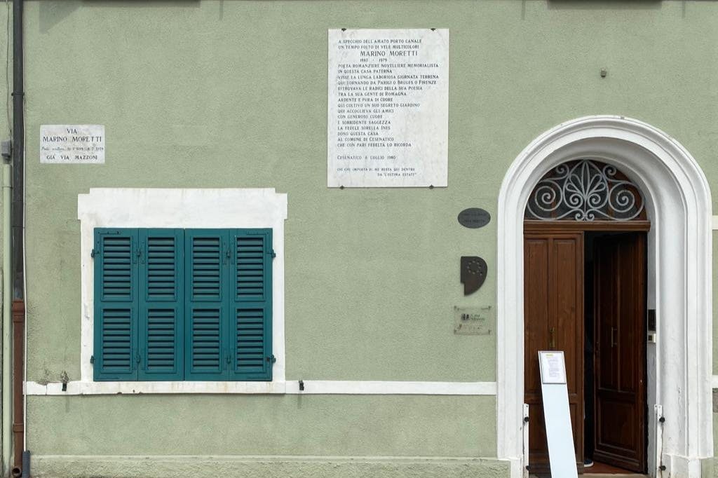 명판, 아치형 나무 문, 청록색 셔터가 있는 녹색 벽. 문자로 된 명판은 문 위와 문 옆에 있습니다.