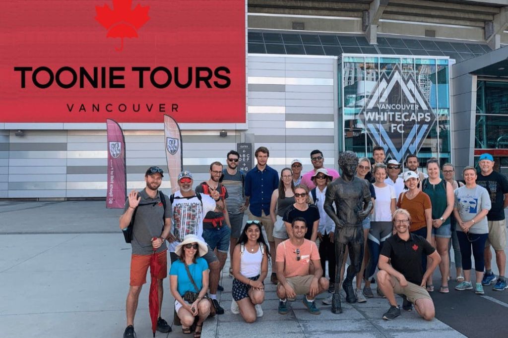 한 무리의 사람들이 도시 분위기의 조각상 주위에서 포즈를 취하고 있습니다. 그 뒤편에는 밴쿠버 화이트캡스 FC 표지판이 보입니다.