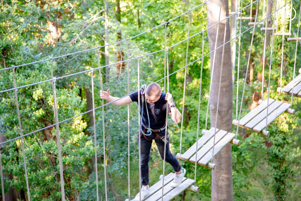 녹색 단풍으로 둘러싸인 숲에서 로프 다리를 탐색하고 안전 하네스를 착용하고 사이드 로프를 잡는 사람.