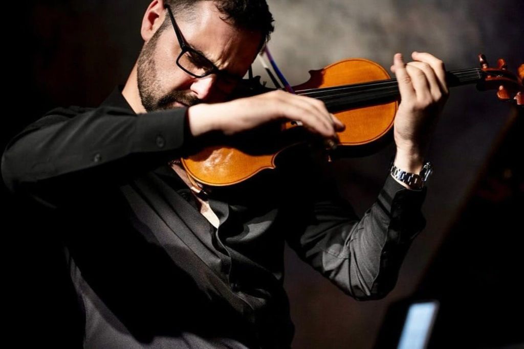 어둡고 흐릿한 배경에 맞춰 초점이 맞춰진 바이올린을 연주하는 안경과 검은 셔츠를 입은 사람.