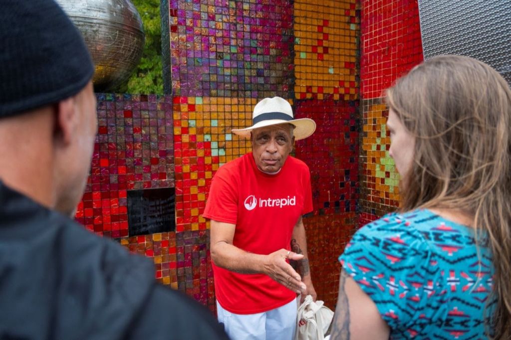 빨간색 '인트레피드 (Intrepid)' 셔츠와 모자를 입은 남성이 화려한 모자이크 벽 앞에서 여성과 대화를 나누고 있습니다.