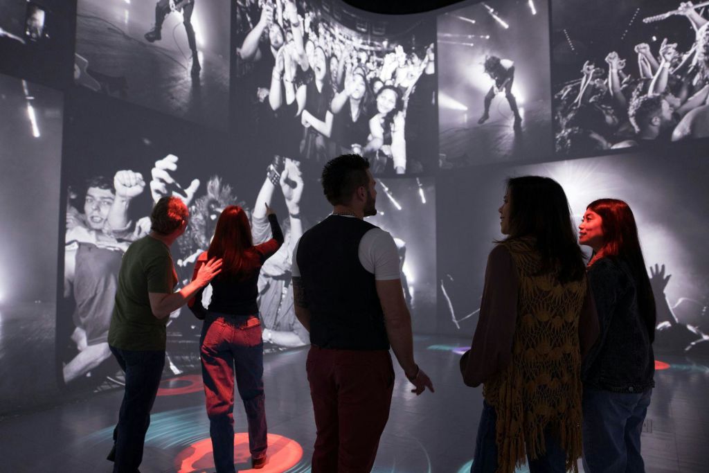 커다란 흑백 콘서트 사진이 벽에 투영된 방에 세 사람이 있습니다.