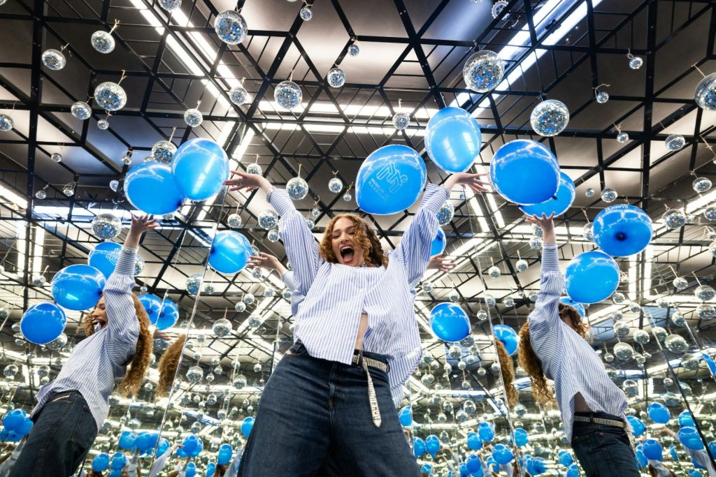 반사되는 표면과 걸이 장식이 있는 거울 방에서 파란 풍선을 들고 즐겁게 포즈를 취하는 사람.
