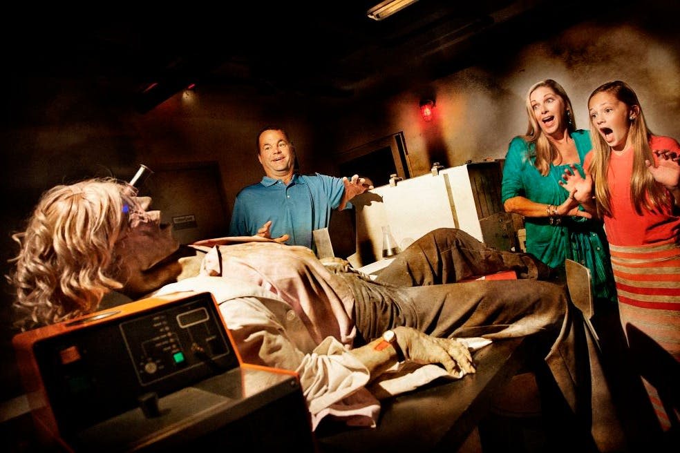 파란색 셔츠를 입은 남성이 실험실 뒤편에 서서 머리카락이 흐트러지고 보철 부상을 입은 앉은 실물 같은 마네킹을 관찰하고 있습니다.