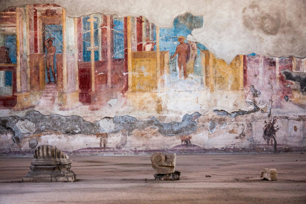 형상과 건축적 요소가 묘사된 화려한 고대 프레스코화가 있는 풍화 벽으로, 돌 조각 조각 조각이 있습니다.