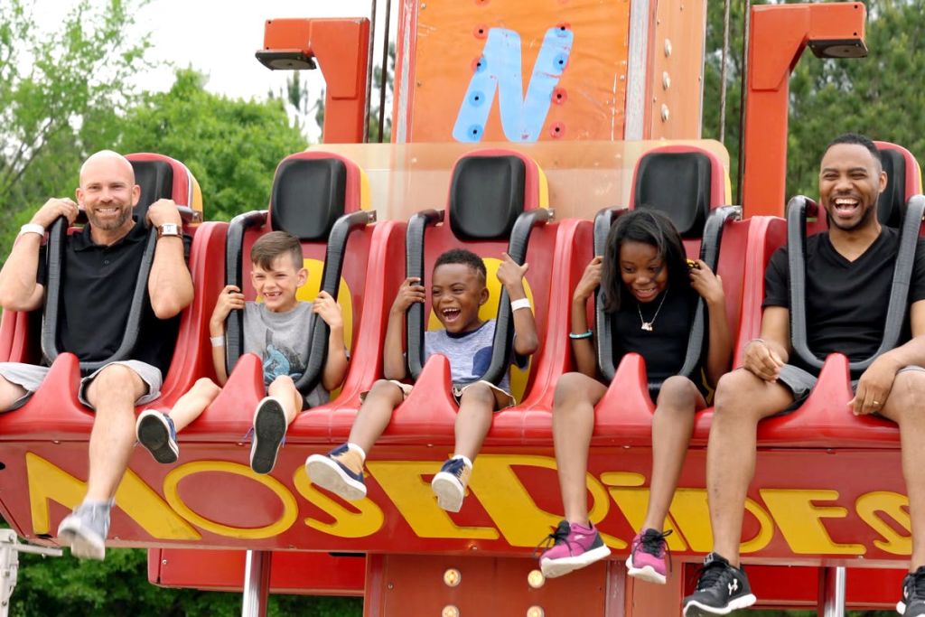 어린이를 포함한 3명이 놀이 기구에 앉아 웃으며 하네스를 붙잡고 있습니다.