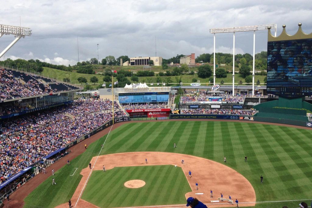 운동장에 선수들이 있고, 스탠드에 관중들이 있으며, 잔디가 깔린 언덕과 건물을 배경으로 한 야구 경기장입니다.