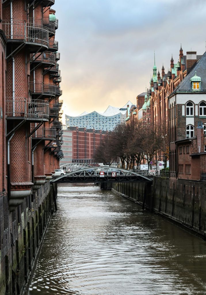 양쪽에 붉은 벽돌 건물이 있는 운하, 가로로 뻗은 금속 다리와 현대적인 유리 건물이 배경입니다.