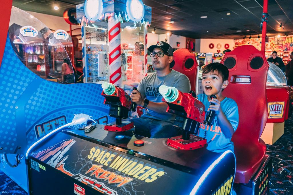 성인 한 명과 어린이 한 명이 밝게 빛나는 아케이드에서 함께 스페이스 인베이더 아케이드 슈팅 게임을 즐기고 있습니다.