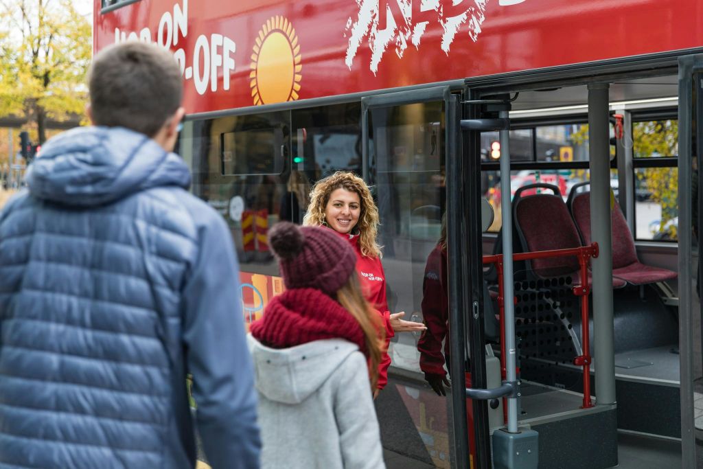 빨간 코트를 입은 미소 짓는 여성이 두 사람을 빨간색 홉온홉오프 투어 버스로 안내합니다.