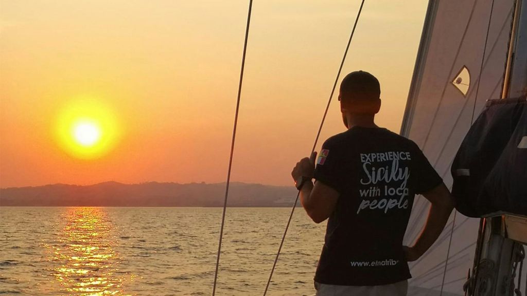 해 질 녘 보트에 올라 밧줄을 잡고 물과 산을 배경으로 '현지인과 함께하는 시칠리아 체험' 셔츠를 입은 사람.