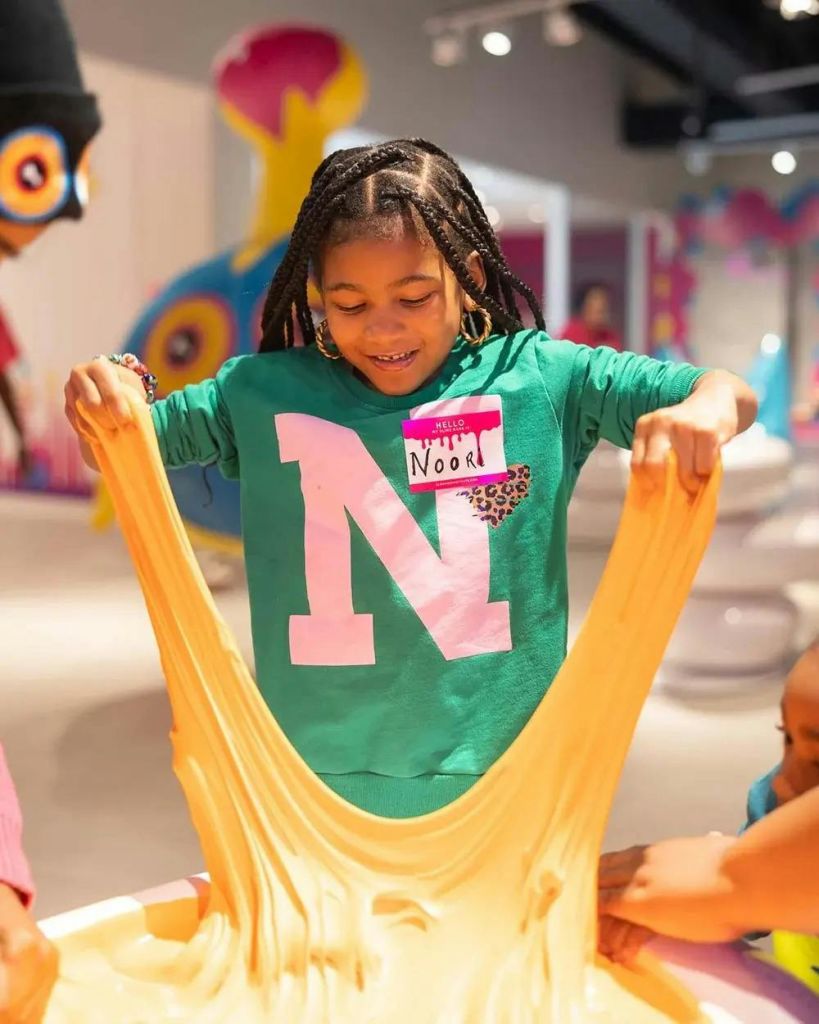 커다란 "N" 과 이름표가 달린 녹색 셔츠를 입은 어린이가 신축성 있는 노란색 물질을 들고 미소를 짓고 있습니다.