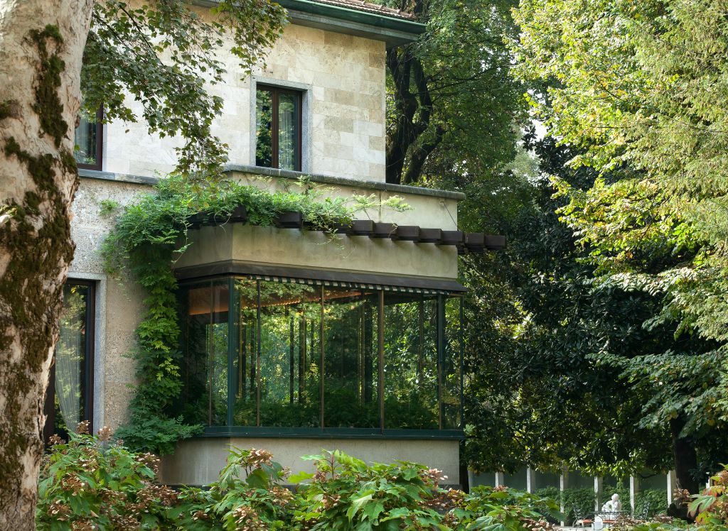 울창한 녹색 식물과 나무로 둘러싸인 커다란 유리로 둘러싸인 일광욕실이 있는 석조 건물입니다.