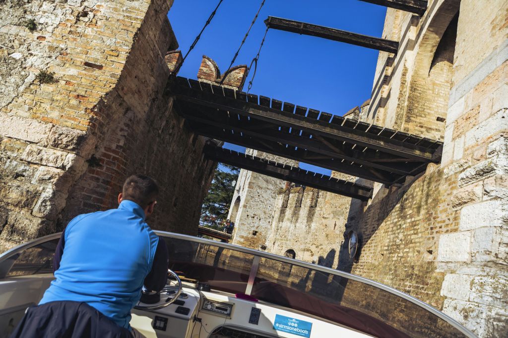 한 사람이 고대 벽돌 벽과 맑고 푸른 하늘로 둘러싸인 쇠사슬이 달린 돌다리 아래에서 모터보트를 타고 항해하고 있습니다.